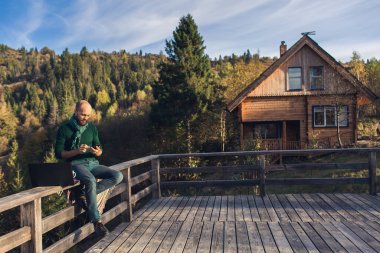 Sakallı adam tırabzanda oturur ve dizüstü bilgisayarda çalışır. Dağlarda tatilde olan serbest yazar hipster..