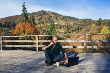 Sakallı adam dağlarda dizüstü bilgisayarla oturmuş telefonla konuşuyor..