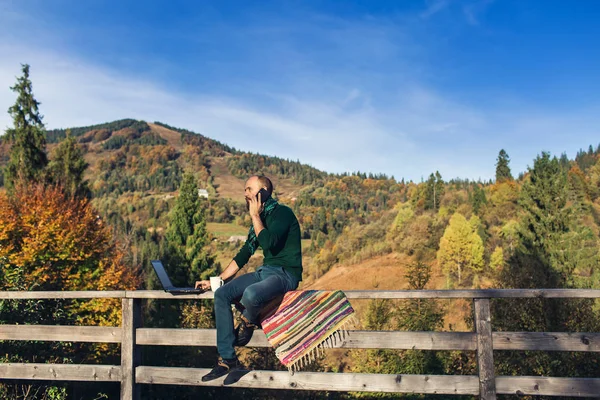 Босоногий мужчина с ноутбуком сидит на перилах и разговаривает по телефону. Хипстер-фрилансер на отдыхе в горах. — стоковое фото