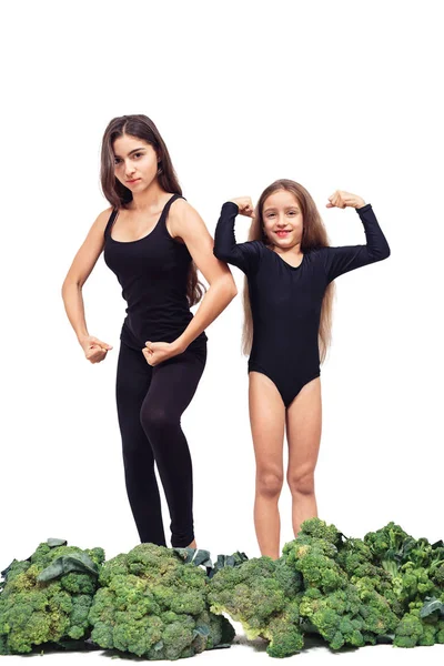Twee meisjes in sportkleding staan tussen broccoli en tonen biceps. — Stockfoto