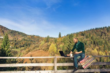 Sakallı adam terasta oturur ve dizüstü bilgisayarla çalışır, telefonda konuşur. Dağlarda tatilde olan serbest çalışan bir hippi.
