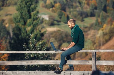Sakallı adam terasta oturur ve dizüstü bilgisayarla çalışır. İnternet üzerinde uzaktan çalışma kavramı