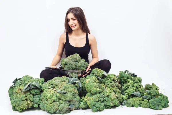 sad tearful girl sitting in big heap of cabbage and holding broccoli in pan. the girl does not like to eat vegetables