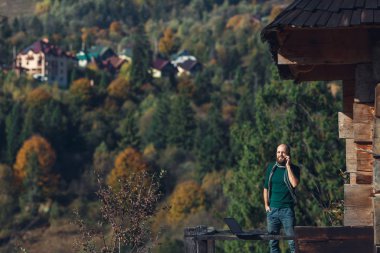 Sakallı adam dizüstü bilgisayarda çalışıyor ve ahşap evin yanındaki terasta telefonla konuşuyor. Bağımsız kavram