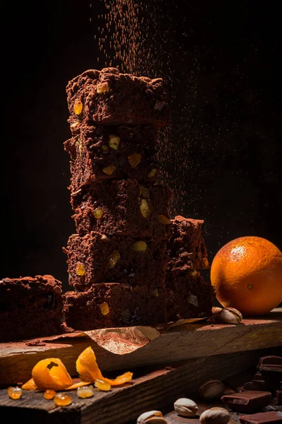 A stack of chocolate brownies on wooden background with orange peels and pistachio nuts, homemade bakery and dessert.