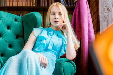 Kameraya bakarak Kütüphanesi'nde sandalyede oturan mavi elbise zarif bayan. Güzellik, moda. Doğal ışık stüdyo.