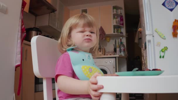 Little two years old blond blue-eyed girl grimaces while having her dining, 4k shot — ストック動画