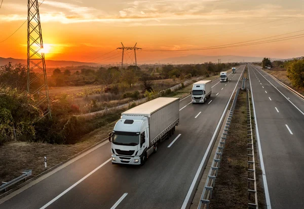 Turuncu gün batımında muhteşem bir gökyüzünün altındaki bir otoyolda mavi kamyon konvoyu. Karavan veya konvoyda beyaz kamyon izleri olan otoyol ulaşımı