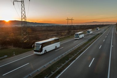 Dört beyaz otobüsün karavanı ya da konvoyu turuncu gün batımında muhteşem bir gökyüzü altında bir otoyolda ilerliyor. Beyaz otobüslü otoyol ulaşımı