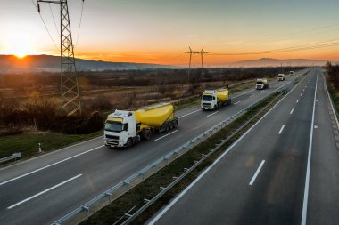 Kırsal alanda, gün batımında, beyaz ve sarı tank kamyonlarıyla dolu bir konvoy ya da karavan.