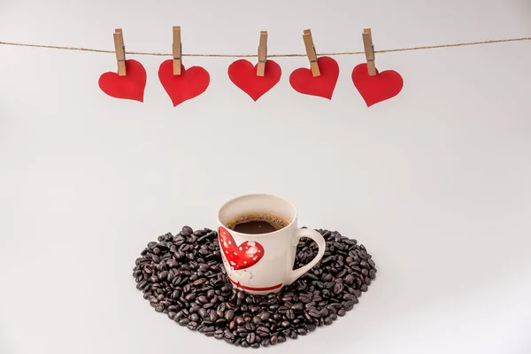 Cup of Coffee with heart made of coffee grains on white background and Clothes pegs and red paper hearts on rope above. Valentines day concept