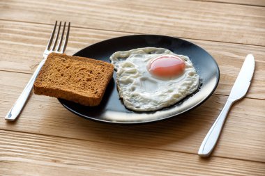 Kızarmış yumurta, ahşap bir masanın üstünde siyah bir tabakta, kızarmış ekmek ve çatal bıçaklarla kaplanmış. Kahvaltılık yemek. Güneşli tarafı yumuşak yağda yumurta