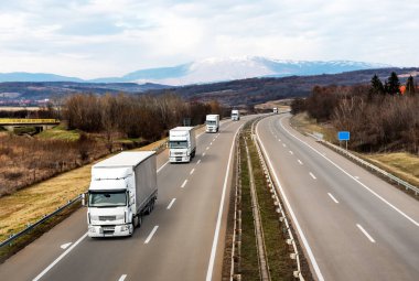 Yoldaki beyaz kamyonlar ya da çekici ekipler konvoyu. Asfalt Otoyolu, Dağların Arkaplanına Karşı Çevre Yolu. İş Ulaşım ve Taşımacılık Endüstrisi.