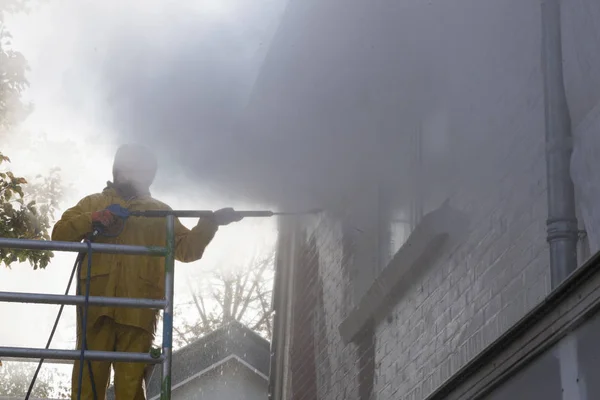 Man in geel regen pak reinigt verf van bakstenen muur voor huis fa — Stockfoto