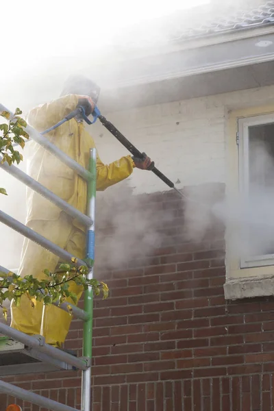 Man in geel regen pak reinigt verf van bakstenen muur voor huis fa — Stockfoto