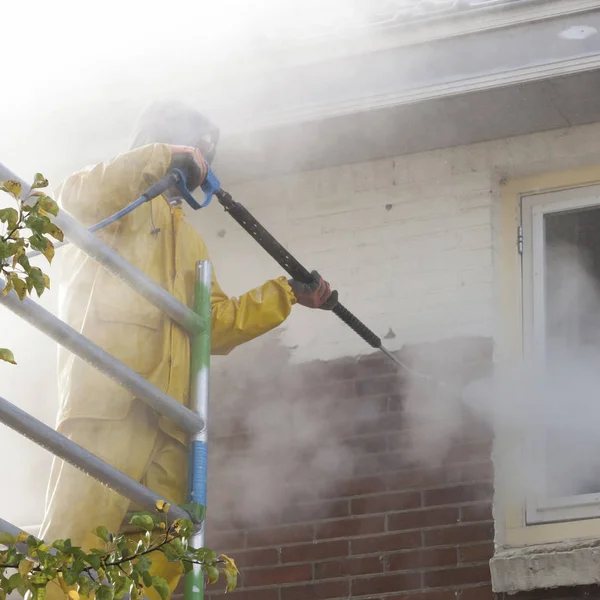 Hombre de traje de lluvia amarillo limpia la pintura de la pared de ladrillo de la casa fa Imagen de stock