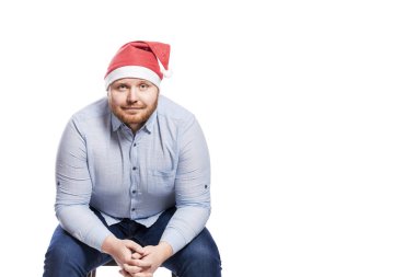 Mavi gömlekli, kot pantolonlu ve Noel Baba şapkalı gülümseyen kızıl saçlı bir adam sandalyede oturuyor. Yakın plan. Beyaz arka planda izole edilmiş. Metin için boşluk.