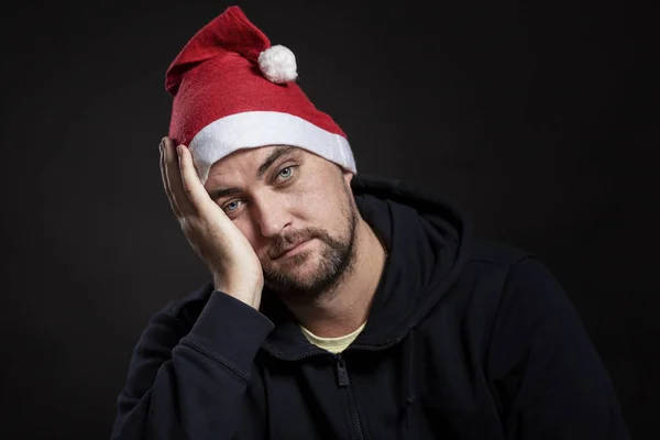 Un homme triste avec une barbe rouge dans un chapeau du Père Noël a couvert son visage de sa main. Fond noir. Gros plan . — Photo