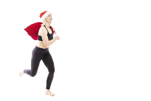 Eine junge lächelnde Frau im schwarzen Trainingsanzug mit Weihnachtsmannmütze und rotem Sack mit Geschenken läuft. volle Höhe. isoliert über weißem Hintergrund. Raum für Text. — Stockfoto