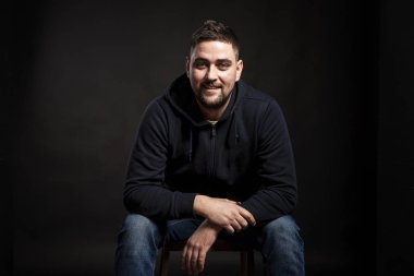 Mavi kapüşonlu genç ve yakışıklı bir adam oturuyor ve kameraya bakıyor. Siyah arkaplan.