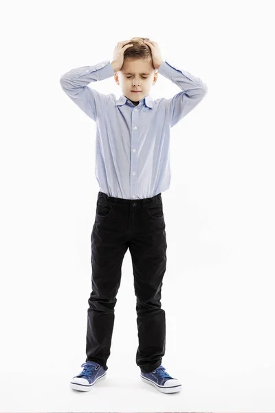 Een leuke serieuze jongen, een 8-jarige schooljongen in een blauw shirt en broek, houdt zijn hoofd in zijn handen. Op volle hoogte. Witte achtergrond. — Stockfoto