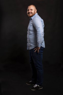 Mavi gömlekli, kot pantolonlu, sakallı, şişman, kızıl saçlı bir adam kameraya bakıp gülüyor. Yan görüş. Siyah arkaplan. 