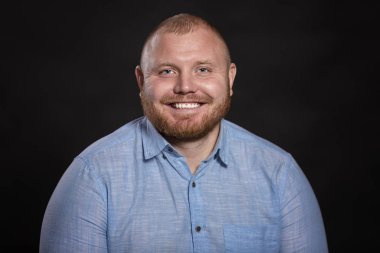 Mavi gömlekli, sakallı, bıyıklı, şişman, kızıl saçlı bir adam gülüyor. Siyah arka plan. Yakın plan..