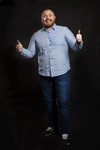 A fat red-haired man with a beard and mustache in a blue shirt and jeans stands holding thumbs up and laughs. Looks at the camera. Black background. — 스톡 사진