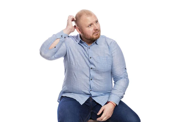 Tlustý zrzavý muž s vousy v modré košili a džínami sedí a škrábe se na hlavě. Izolované přes bílé pozadí. — Stock fotografie