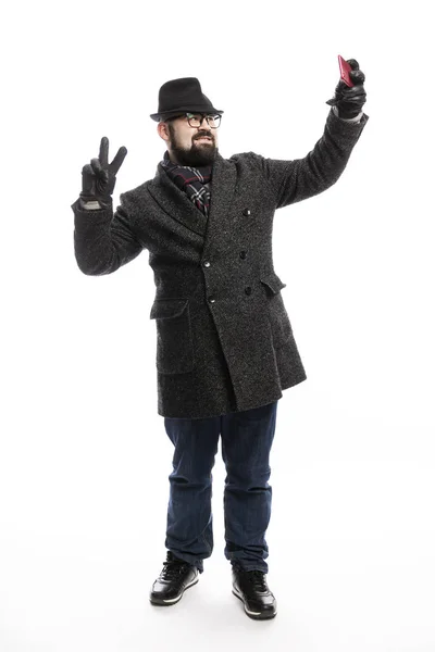 Elegante hombre con barba, vestido con un elegante abrigo y sombrero hace selfie. A toda altura. Aislado sobre fondo blanco . — Foto de Stock