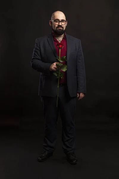 Łysy mężczyzna w okularach i broda w garniturze z czerwoną różą w ręku. Czarne tło. — Zdjęcie stockowe