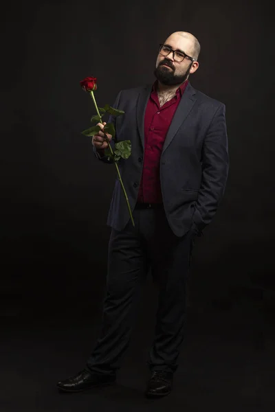 Łysy mężczyzna w okularach i broda w garniturze z czerwoną różą w ręku. Czarne tło. — Zdjęcie stockowe