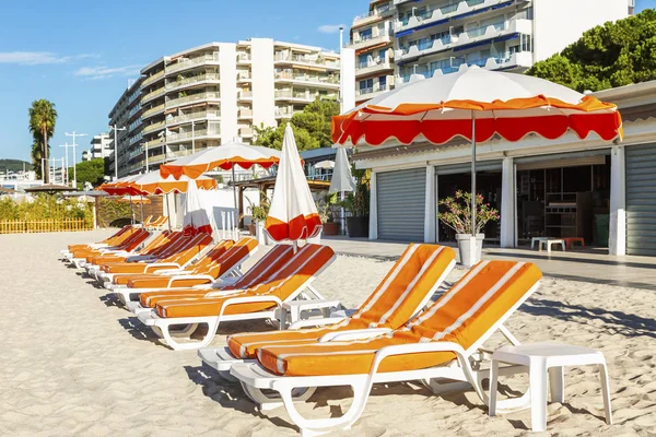 A row of deck chairs with orange mattresses on a sandy beach in a resort town. Bright sunny day.