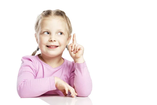Een klein meisje in een roze trui zit aan tafel en steekt haar wijsvinger op. Geïsoleerd op witte achtergrond. Ruimte voor tekst. — Stockfoto
