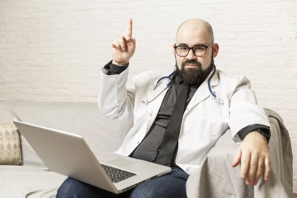 一名男医生坐在沙发上 在笔记本电脑前工作 远程工作 流行病期间的在线咨询 — 图库照片