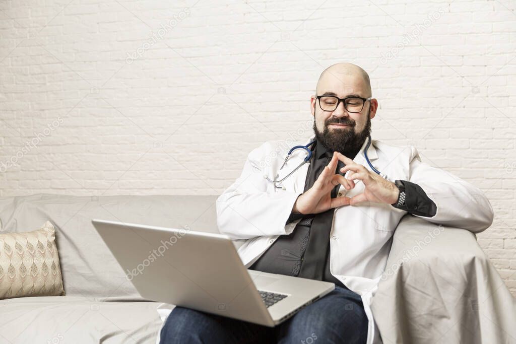 A male doctor sits on a sofa and works in front of a laptop. Remote work, online consultations during the epidemic.