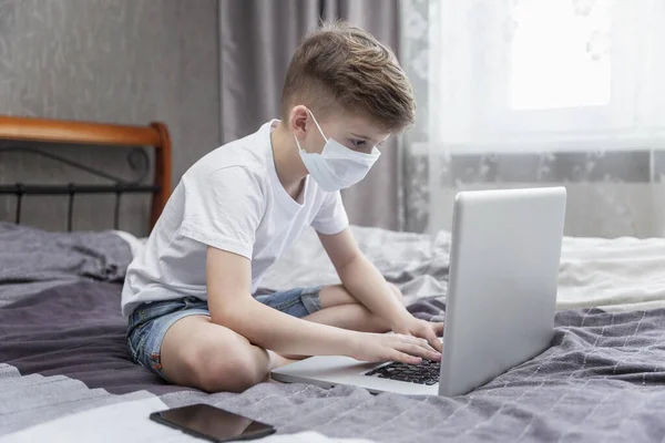 Tıbbi Maskeli Bir Çocuk Evde Yatağında Dizüstü Bilgisayarın Önünde Oturuyor — Stok fotoğraf