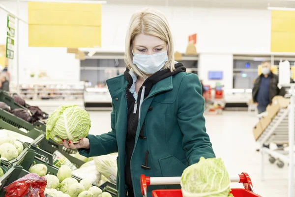 Tıbbi maskeli sarışın kadın sebze bölümündeki bir süpermarketten alışveriş yapıyor. Koronavirüs salgını sırasında karantina.
