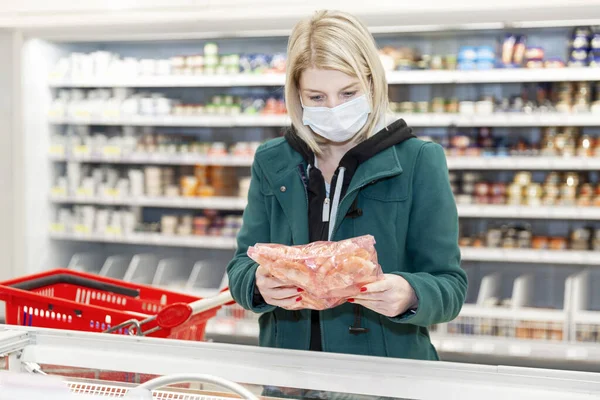 Tıbbi maskeli sarışın bir kadın dondurulmuş gıda bölümündeki süpermarketten alışveriş yapıyor. Koronavirüs salgını sırasında karantina.