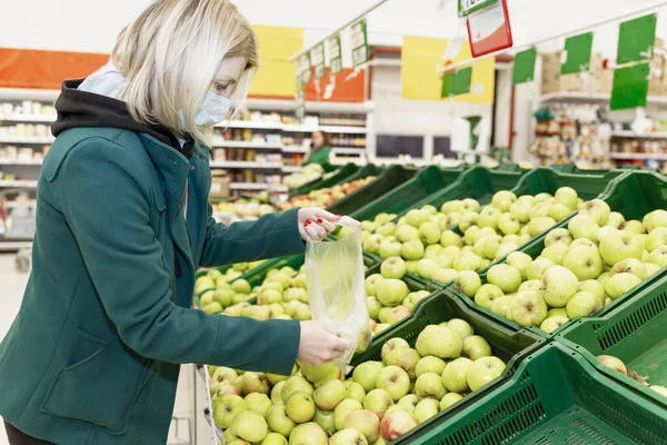 Woman blonde in a medical mask chooses vegetables in a supermarket. Healthy eating and vegetarianism. Quarantine during the coronavirus pandemic.