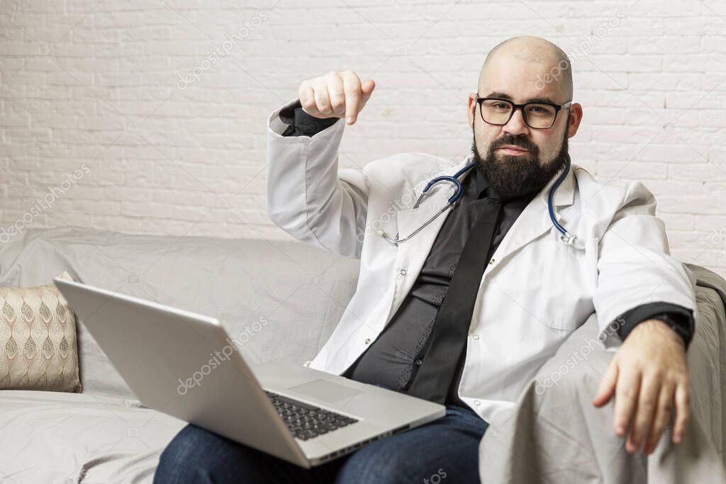 A bald male doctor in a white coat with glasses sits with a laptop on the couch. Online consultations. Quarantine during the coronavirus pandemic.