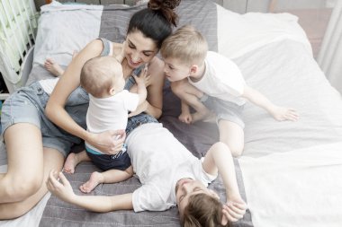 Küçük oğullarıyla birlikte genç bir anne yatakta uzanıyor ve gülüyor. Aile ilişkilerinde şefkat. Koronavirüs salgını sırasında karantina.