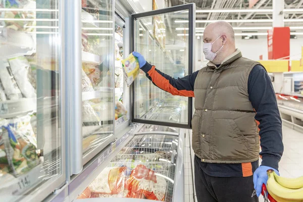 A middle-aged man in a medical mask and gloves chooses frozen foods in a supermarket refrigerator. Healthy eating during the coronavirus pandemic.