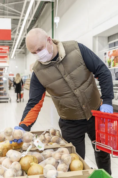 Bald adult male in medical mask and gloves chooses vegetables in a supermarket. Self-isolation regime during the coronavirus pandemic. Vertical.