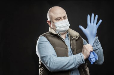 Kel ve maskeli bir adam ellerine mavi tıbbi eldiven takıyor. Koronavirüs salgını sırasında alınan önlemler. Siyah arkaplan.