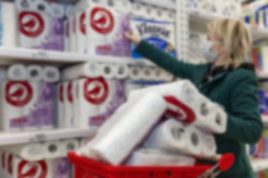 A blond woman in a medical mask buys a lot of toilet paper in a supermarket. Panic and deficiency during the coronavirus pandemic. Blurred. clipart