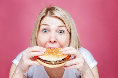 Sarışın kadın büyük sulu bir hamburger yiyor. Abur cubur ve sağlıksız bir yaşam tarzı. Pembe arka plan. Yakın plan..