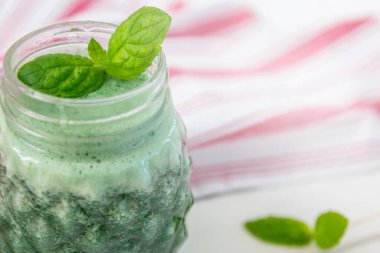 Kavanozda nane yapraklarıyla süslenmiş spirulinalı yeşil smoothie. Arka planda kırmızı çizgili bir mutfak havlusu. Sağlıklı yaşam tarzı.