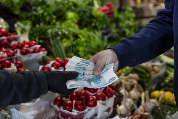 Money in hand against the background of the counter with vegetables at the market. A man pays in rubles. Close-up. The high cost of fresh food.