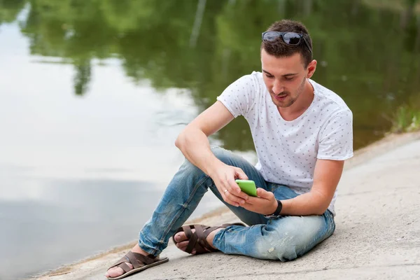 男に携帯電話で見えます。湖のほとりに座っています。. — ストック写真